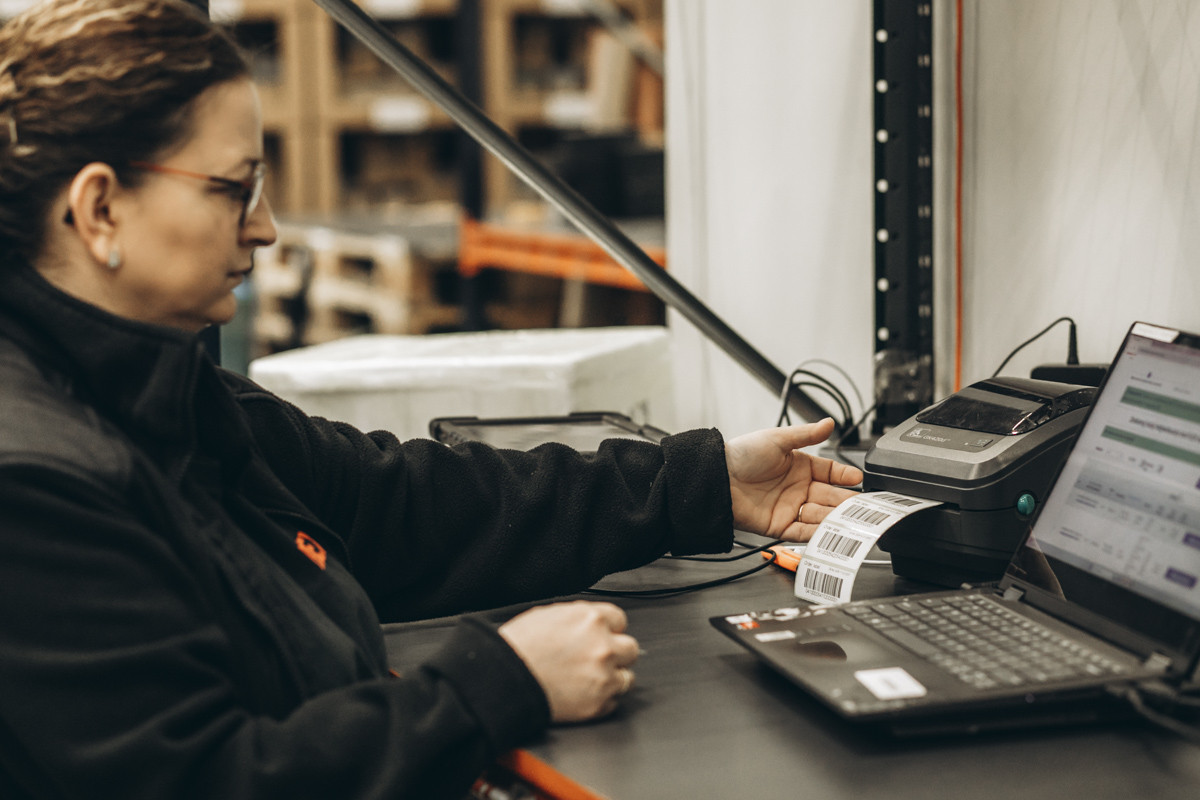 Warehouse employee printing shipping labels at a workstation with a laptop and thermal label printer in a fulfillment facility.
